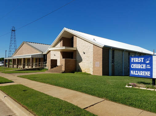 A church building with a brick exterior, white metal roof, and green lawn. A blue sign in front reads 